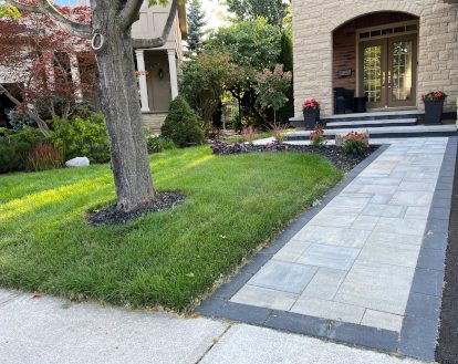 Standard sized front walkway consisting of square and rectangular paver stones in varying colours beside a paved driveway, exemplifying Interlocking Driveway Extensions work on a medium-sized area