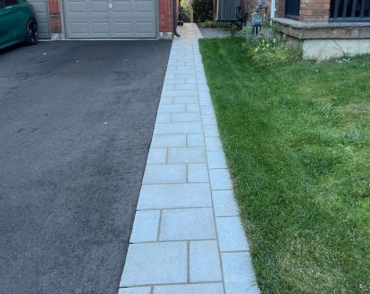 Narrow front walkway consisting of light grey square and rectangular paver stones beside a paved driveway, exemplifying Interlocking Driveway Extensions work on a small area