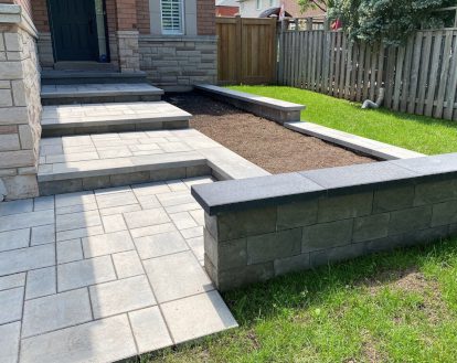 Intricate paver stone design consisting of large grey pavers of varying sizes on a front walkway consisting of 4 large step up platforms beside a long garden bed, exemplifying Interlocking paver work on a large front entrance