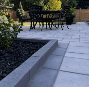 Instagram image post thumbnail showing closeup of grey paver stone patio with different sizes and shades of stones and a stone curb around a bed of black mulch with flowers.