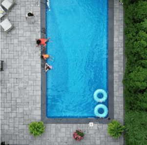 Instagram image post thumbnail showing aerial view of large grey paver stone patio with planters and chairs around an in-ground pool with three children playing in it.