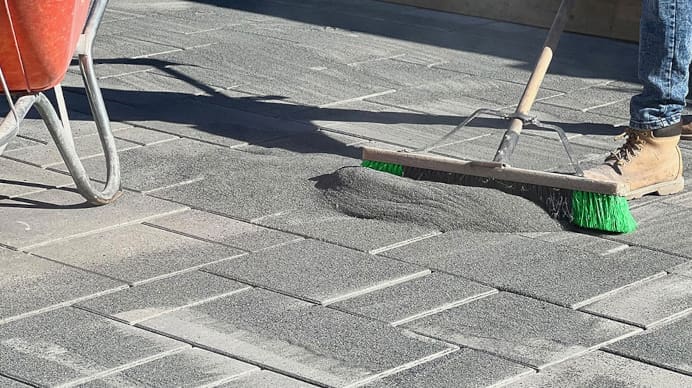 Polymeric sand being pushed across a paver stone patio with a large broom being held by an individual who is mostly cropped out of the image besides his/her right pant leg and boot.