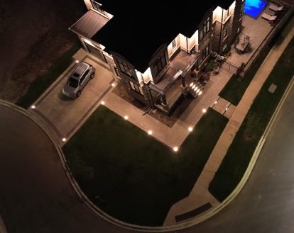 Aerial view of a property at night with recessed lights along paver stone driveway and walkway, as well as front porch steps, exemplifying Landscape Lighting work in a large area