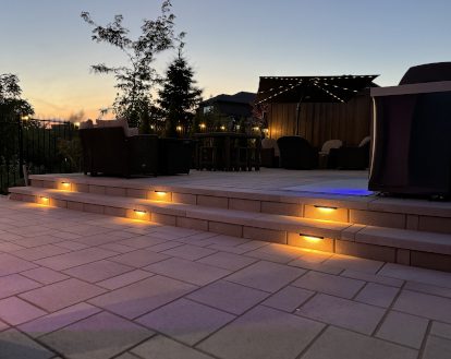 Residential backyard with large paver stone patio consisting of 2 levels with 2 steps that are each lit up 3 small recessed warm yellow lights, exemplifying landscape lighting work in a medium-sized area