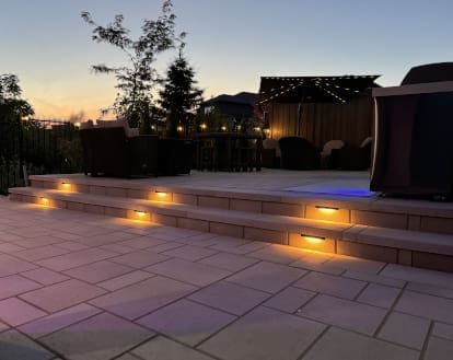 Residential backyard with large paver stone patio consisting of 2 levels with 2 steps that are each lit up 3 small recessed warm yellow lights, exemplifying landscape lighting work in a medium-sized area