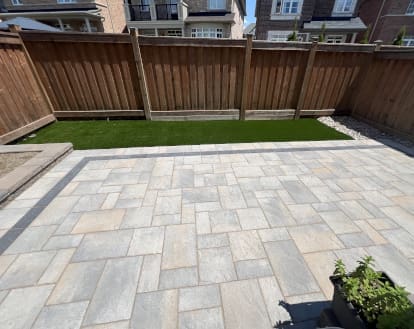 Small rectangular patch of artificial turf between paver stone patio and wooden fence in a residential backyard, exemplifying artificial turf work on a small area