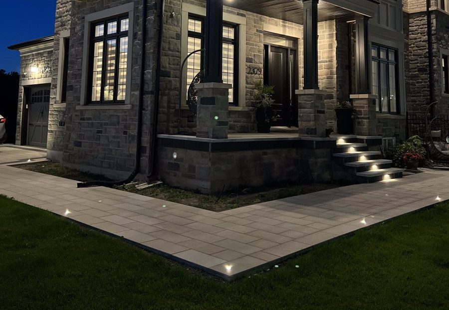 Grey paver stone walkway with recessed lights wrapping the front and left side of a large residential property and a paver stone porch with paver stone steps with recessed lights in the middle of each riser.