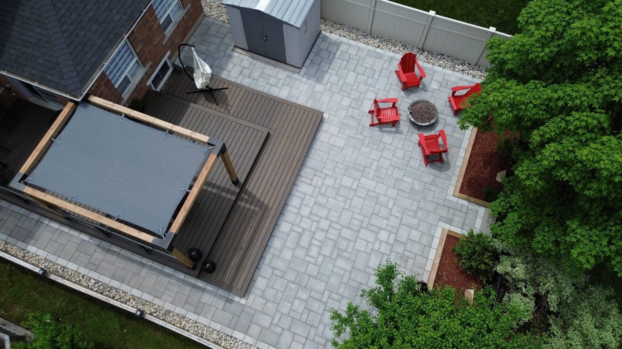 Aerial view of patio with grey rectangular interlocking stones of various sizes. The patio is furnished with a raised gazebo and red muskoka chairs around a fire pit and has a river stone garden around its perimeter.