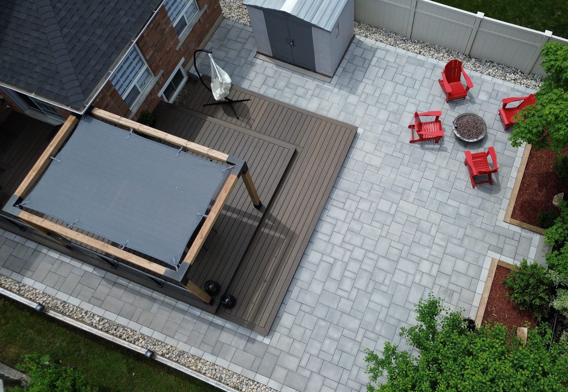 Aerial view of a backyard space with a large grey interlocking stone patio with a raised gazebo on one end and a small fire pit surrounded by 4 red Muskoka chairs on the other end.