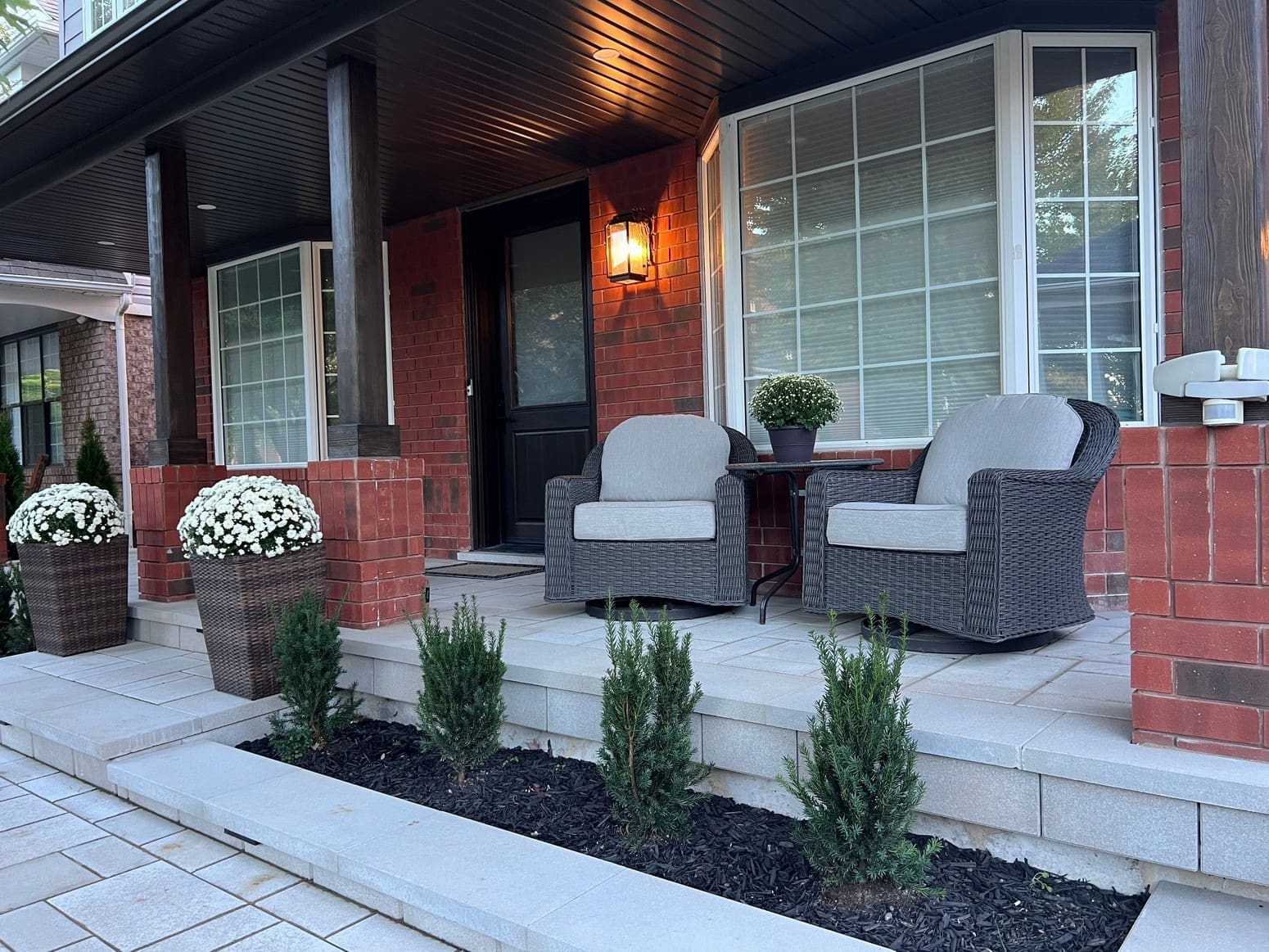A 2 step front porch made with large grey paver stones and a matching paver stone front walkway and curb around a garden with black mulch and baby cedars