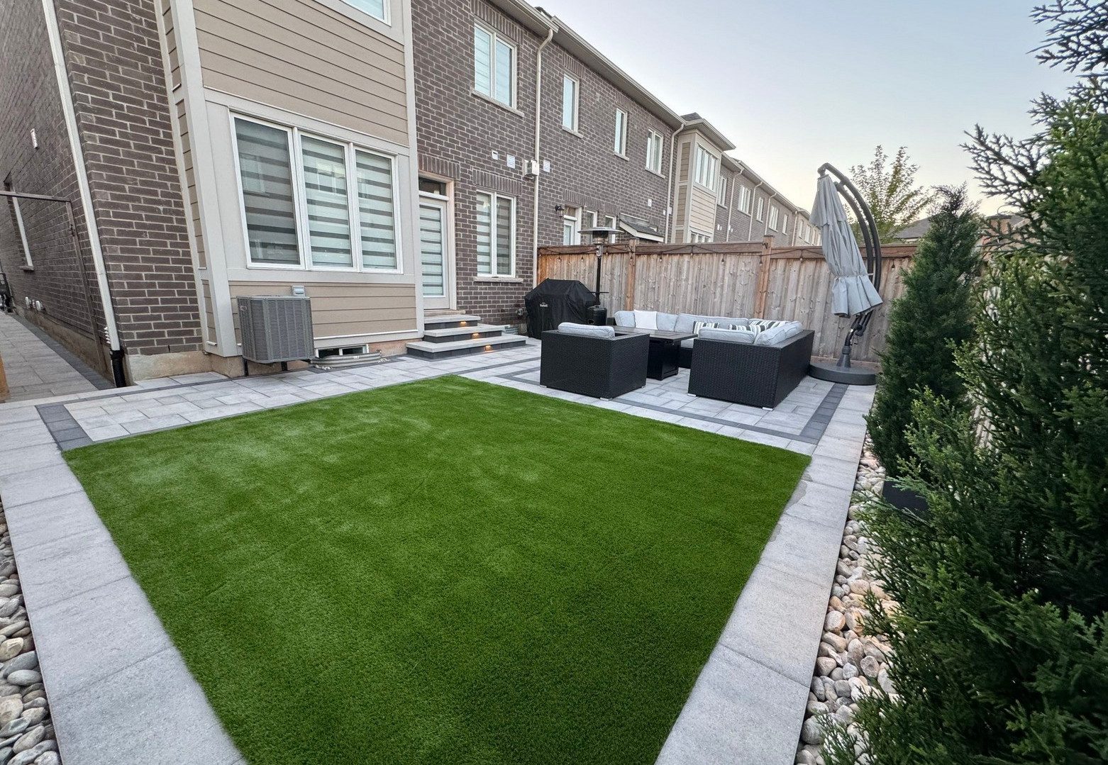 Bright green square turf section with a grey interlocking paver stone border around its perimeter and a grey interlocking stone patio furnished with modular wicker furniture in the background.