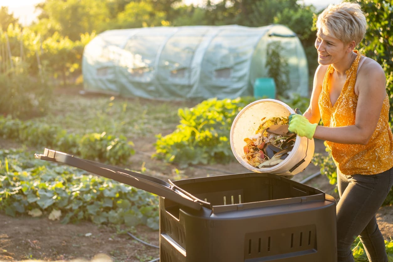 Woman adding kitchen scraps to a backyard compost bin in a landscaped vegetable garden.