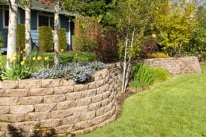 Curved stone retaining wall with built-in flower beds, colorful perennials, and trees in a well-maintained residential backyard.