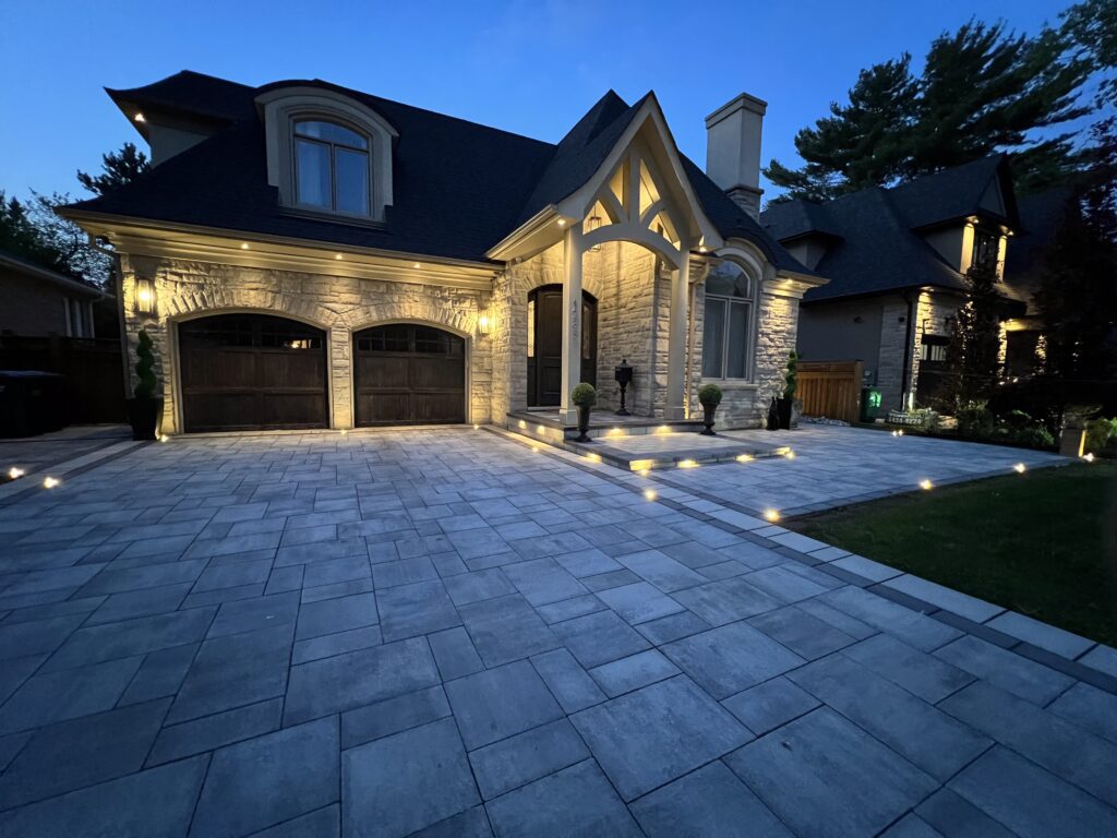 A gray stone driveway made of interlocking pavers features a geometric pattern in front of a modern home, bordered by stone steps and landscaping.