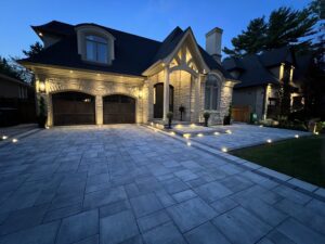 A gray stone driveway made of interlocking pavers features a geometric pattern in front of a modern home, bordered by stone steps and landscaping.