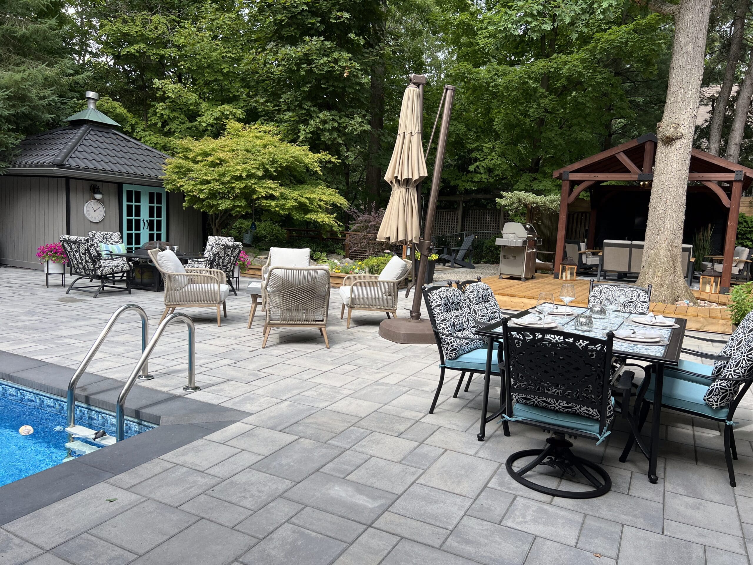 Outdoor interlock patio with green cushioned chairs and a round table beside a swimming pool, shaded by a large umbrella in a fenced backyard.