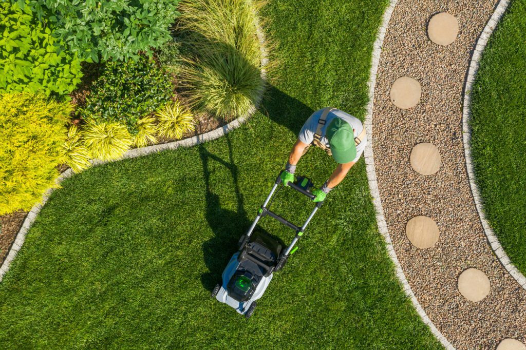 Top-down view of a person mowing a green lawn bordered by decorative landscaping and curved interlocking stone pathway.