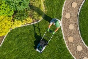 Top-down view of a person mowing a green lawn bordered by decorative landscaping and curved interlocking stone pathway.