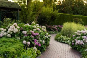 Curved brick interlocking walkway lined with lush greenery and blooming hydrangeas in shades of purple and white, leading through a landscaped garden with a wooden gazebo on the left.