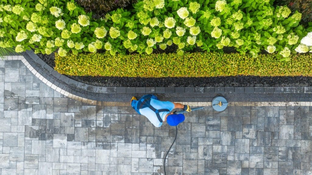 Professional cleaning interlock driveway with power washer beside landscaped garden.