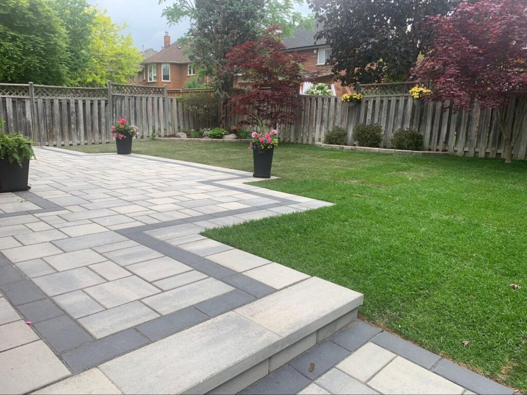 Backyard interlocking patio walkway with planters and lush lawn, surrounded by mature trees and seasonal landscaping.