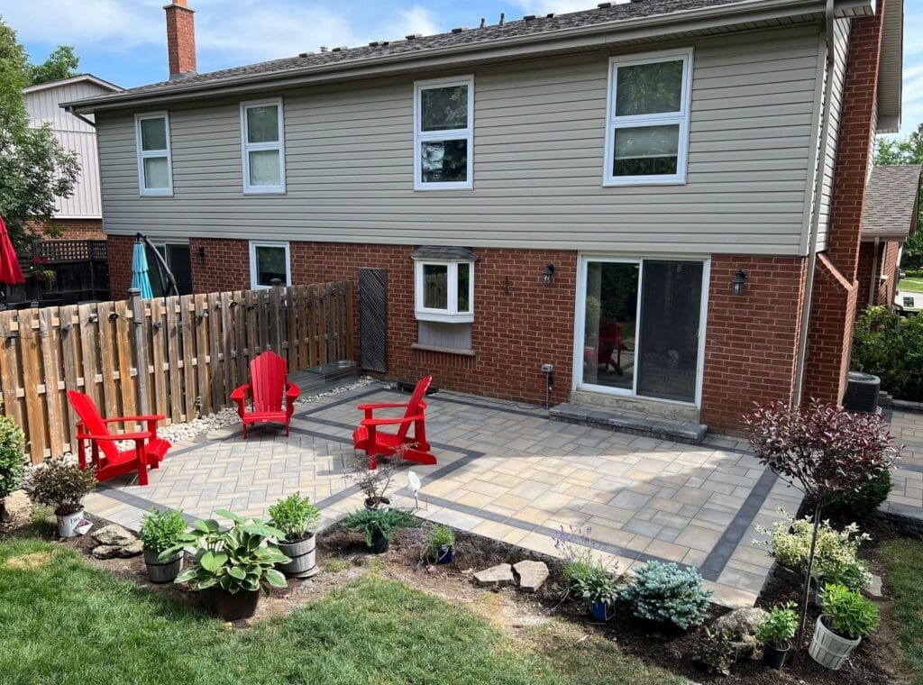Backyard stone patio with a herringbone interlock design, bright red chairs and surrounding garden beds in a landscaped suburban outdoor space.