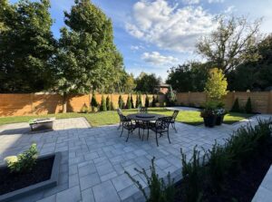 Large backyard featuring a clean interlock patio, circular dining set, structured greenery and privacy fencing, an example of modern landscaping design.
