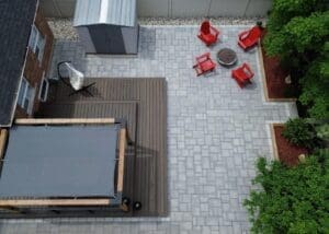 Aerial view of a modern interlock patio with light gray stone pavers, red Adirondack chairs around a fire pit and integrated deck space in a suburban backyard.