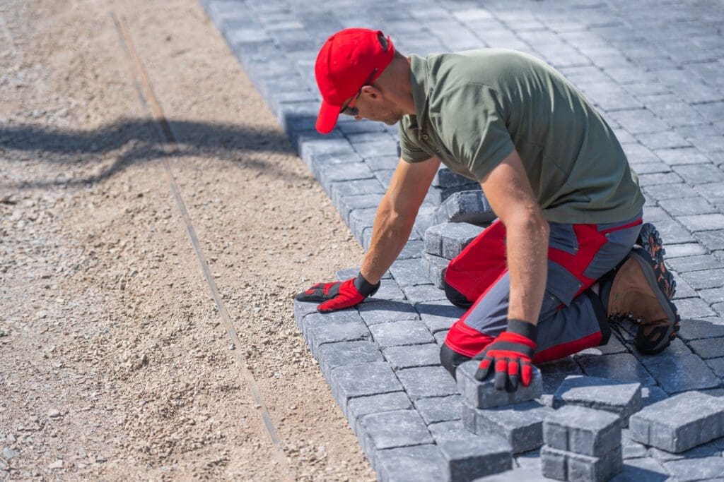 Professional landscaper installing interlocking pavers on a gravel base, wearing red gloves and protective gear.