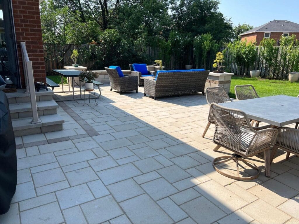 Spacious backyard patio with light interlocking stones, outdoor dining area and modern wicker furniture surrounded by a green lawn.
