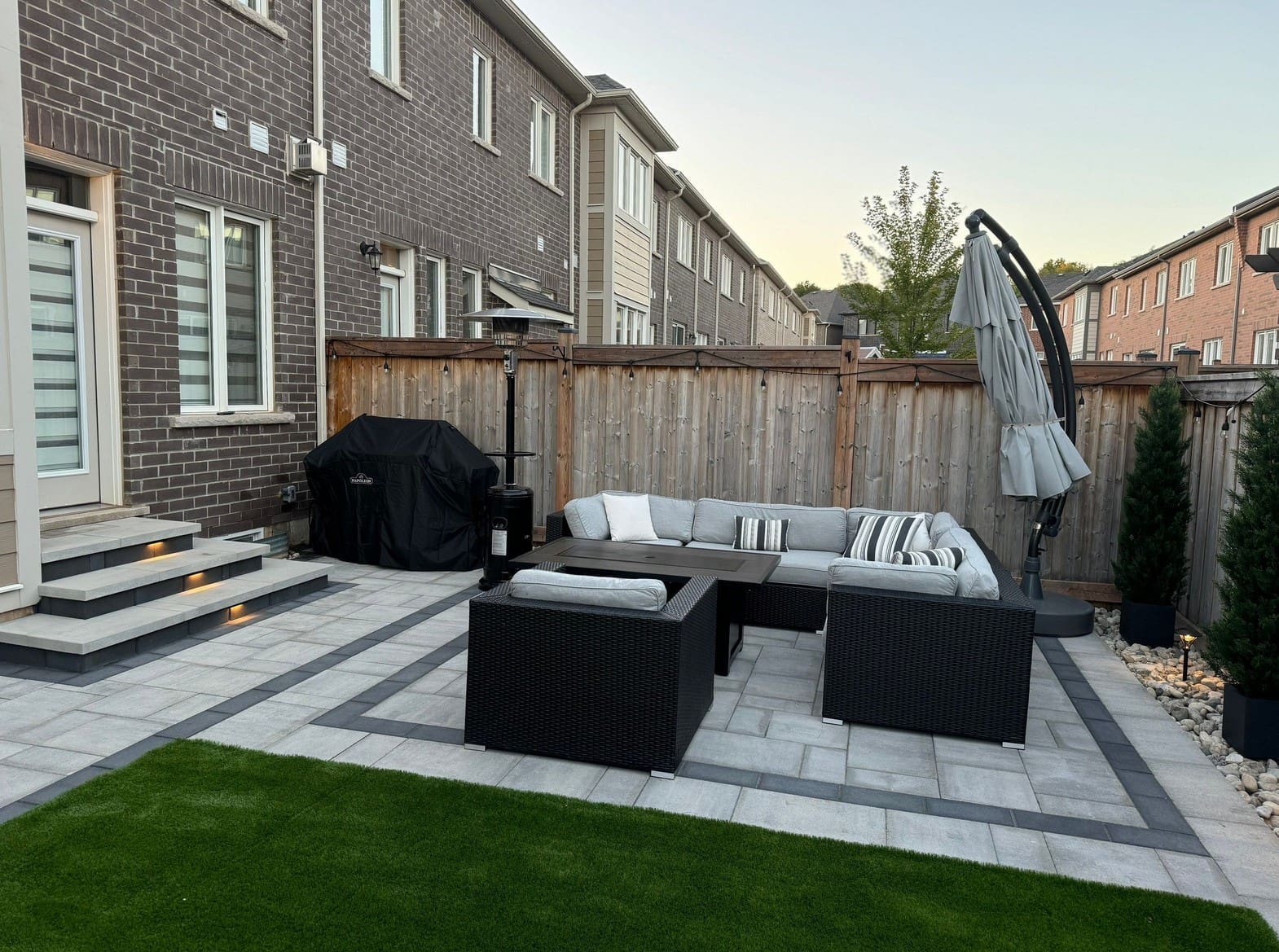 Backyard interlocking design with outdoor furniture, a covered lounge area and subtle landscaping around a spacious patio.