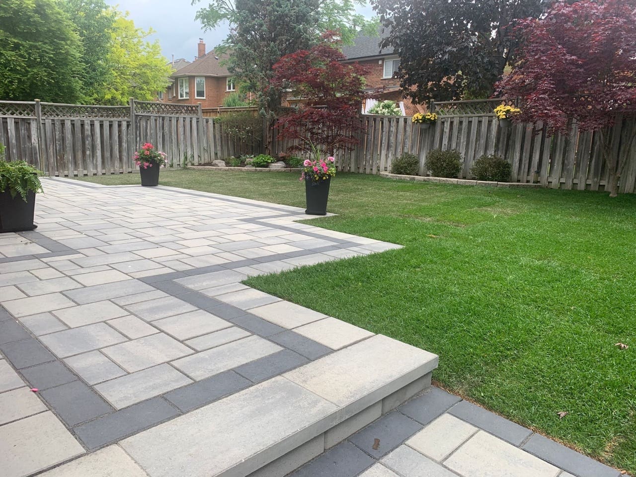 Close-up of backyard interlock design with contrasting dark borders, raised steps, potted flowers and mature trees.